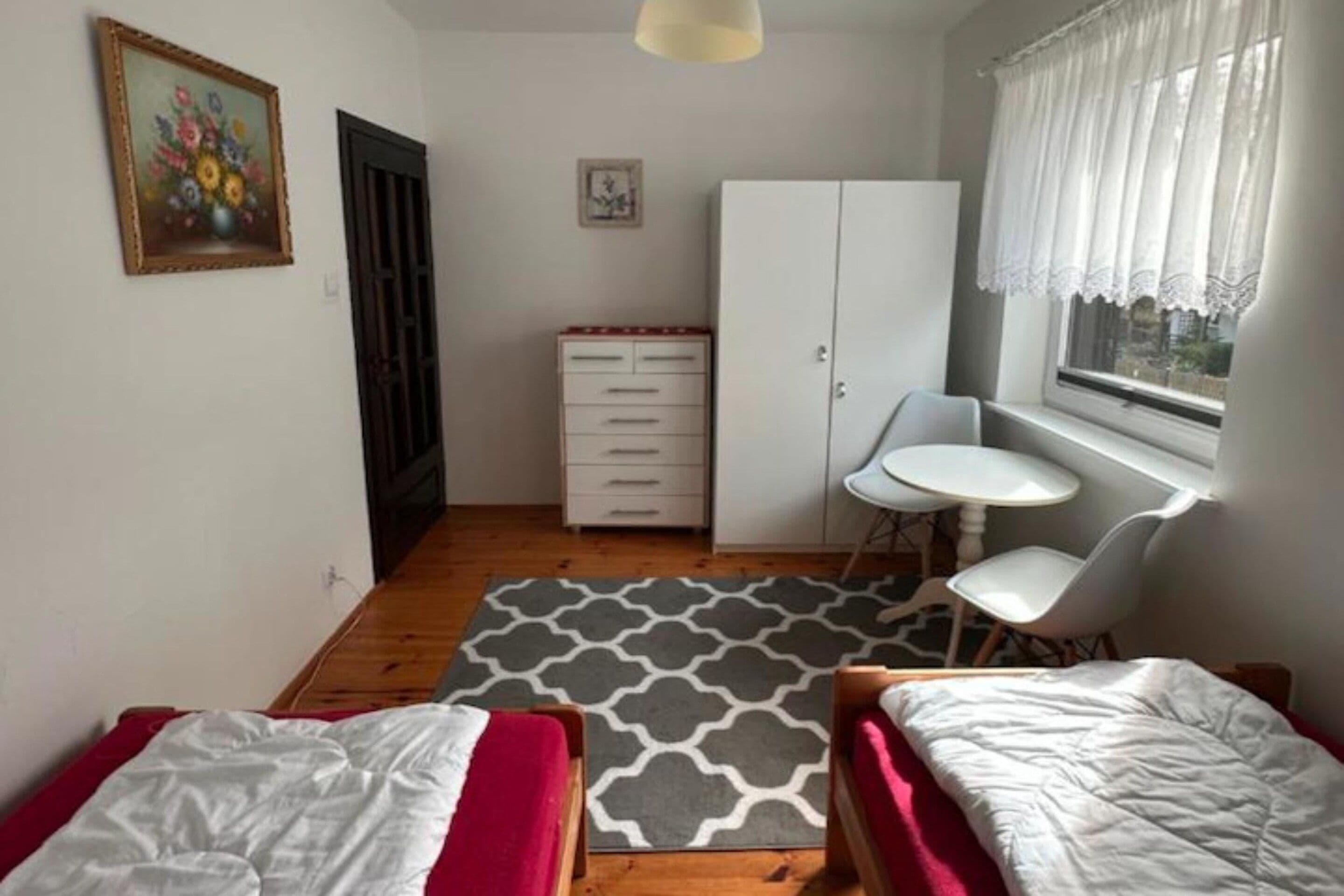 Quarto com duas camas, uma escrivaninha pequena, uma cadeira, guarda-roupa e janela com cortinas.