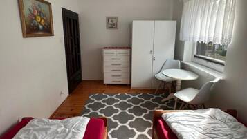 Quarto com duas camas, uma escrivaninha pequena, uma cadeira, guarda-roupa e janela com cortinas.