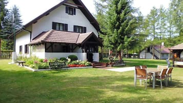 Uma casa de dois andares com telhado inclinado, varanda frontal e gramado bem cuidado, com um banco e uma mesa.