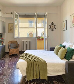 Ein Hotelzimmer mit einem Bett, einem Korbstuhl, einer Heizung, einer Uhr und Blick auf Gebäude durch offene Türen.