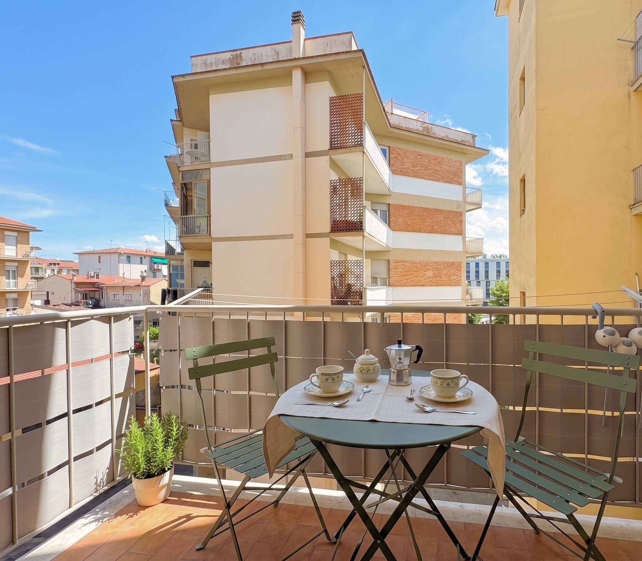 A balcony with a table set for two, chairs, a potted plant, and a view of neighboring buildings.