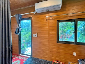 A room with wooden walls, a window with a view of greenery, an air conditioning unit, and a curtain.