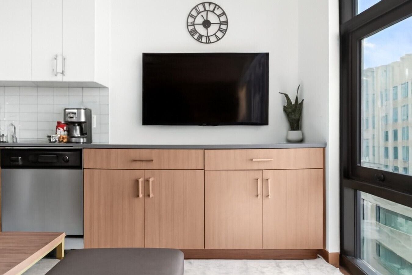 A modern kitchen with a flat-screen TV, wooden cabinets, a coffee machine, and a window with a view of buildings.