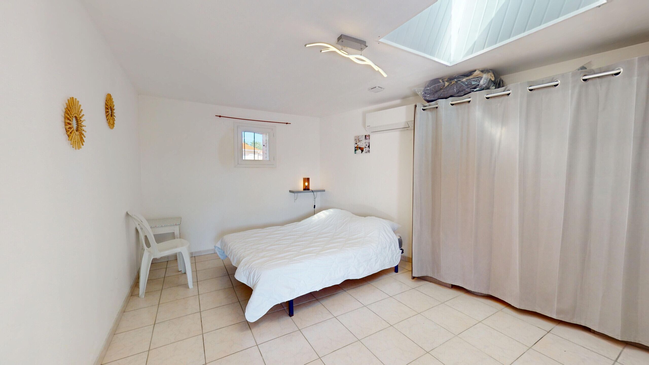 Un dormitorio con cama, una silla, un ventanal pequeño, una lámpara en el techo y una cortina.