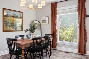 Une salle à manger avec une table en bois et des chaises noires, un lustre et une grande fenêtre avec des rideaux.