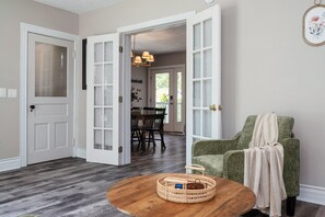 Een kamer met een houten vloer, een groene fauteuil, een ronde salontafel en een eethoek met een tafel en stoelen.