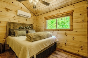 A wooden cabin bedroom with a bed, pillows, a window, and an air conditioning unit. - 10 Minutes from downtown. Valley Hideaway Cottage (Sevierville)