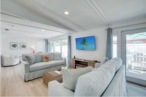 A spacious living area with a light-colored sofa, a coffee table, and a television. There are framed pictures on the wall and a large window with curtains. - Misty Beach, hosted by Beach Retreats on AMI (Bradenton Beach)