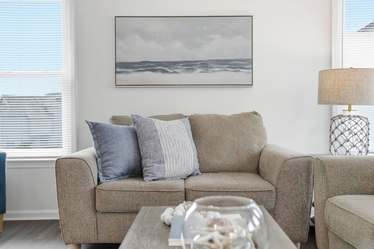 A living room with a beige sofa, a framed picture of the ocean, a lamp, and a glass vase on a coffee table.