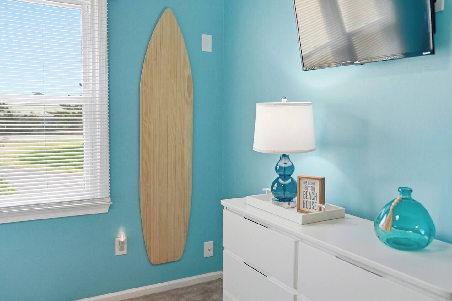 A room with a surfboard on the wall, a lamp, a framed sign, and a vase on a dresser.