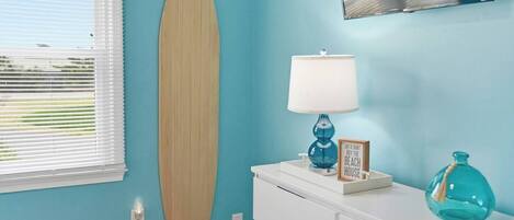 A room with a surfboard on the wall, a lamp, a framed sign, and a vase on a dresser.