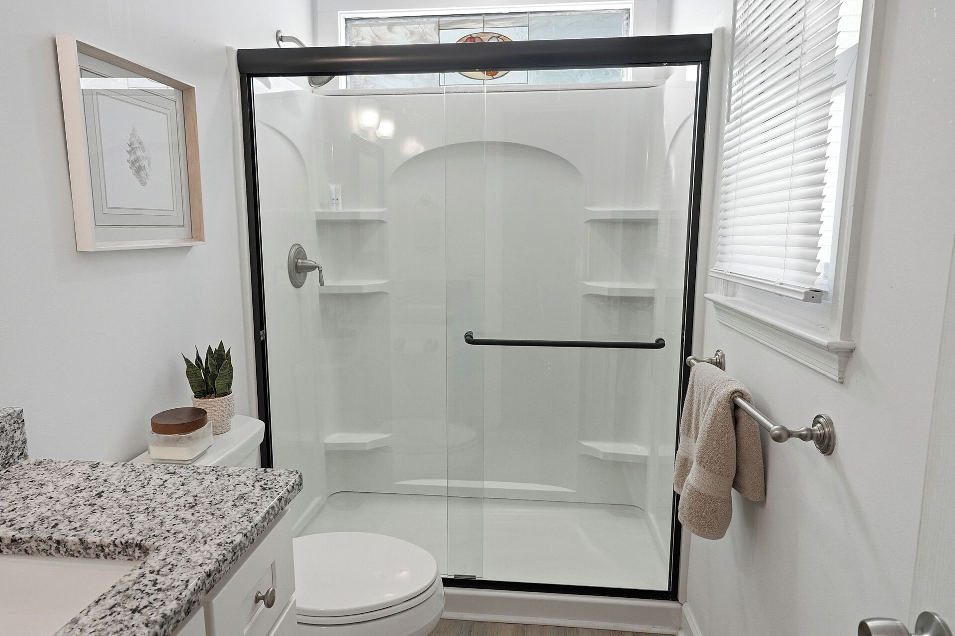 Un baño moderno con ducha envuelta en vidrio, inodoro, lavamanos con encimera de granito y toallero.
