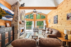 A cozy cabin interior with a stone fireplace, wooden walls, a sofa, and a large window offering a view of the outdoors. - Cedar Ridge Haven | Hot Tub, Fireplace & More! (Gatlinburg)