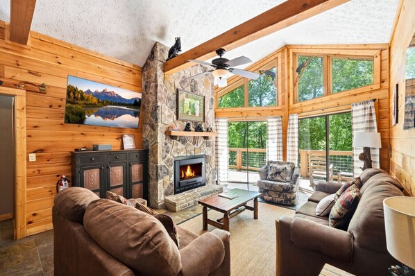 A cozy cabin interior with a stone fireplace, wooden walls, a large window, and a comfortable seating area. - Cedar Ridge Haven | Hot Tub, Fireplace & More! (Gatlinburg)