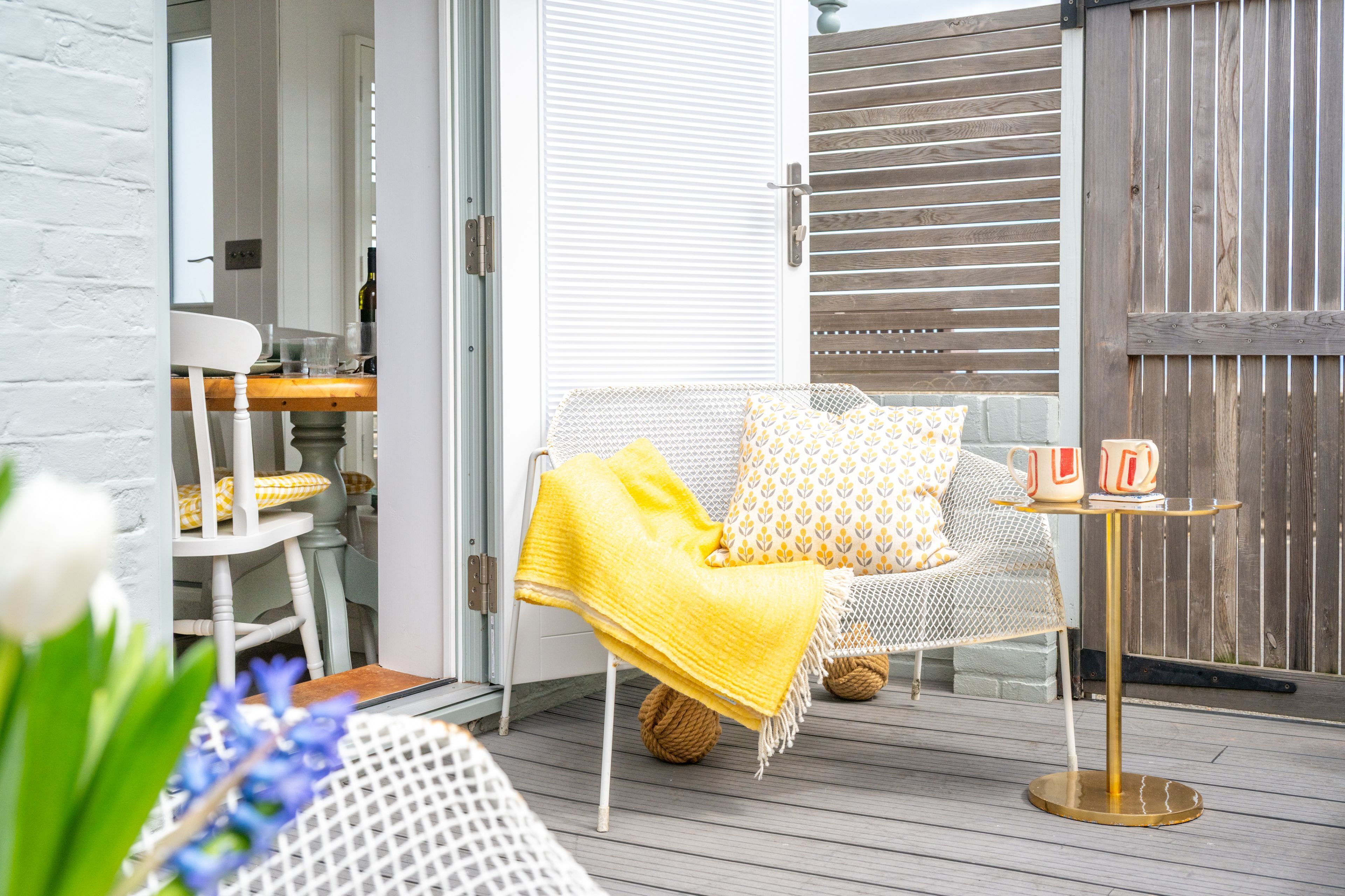 En terrass med en vit stol, en soffa med mönstrade kuddar och ett litet bord med koppar.