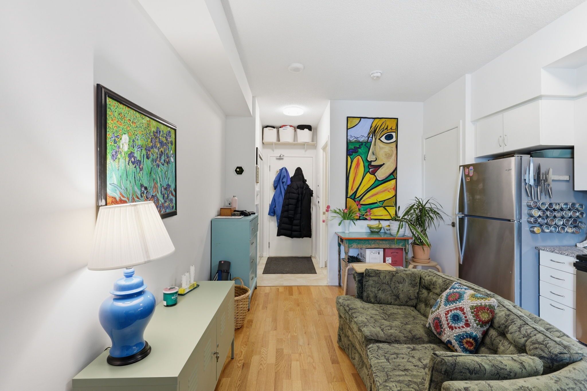 Un espacio habitable pequeño con kitchenette, un sofá con un cojín colorido, una lámpara azul y un cuadro en la pared.