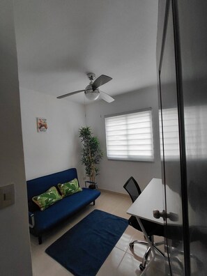 Een kamer met een plafondventilator, een bank met kussens, een kleine tafel, een potplant en een raam met jaloezieën.