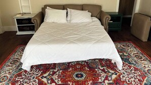 Una cama con un edredón blanco, una alfombra estampada, un sofá marrón y una mesita con un libro.