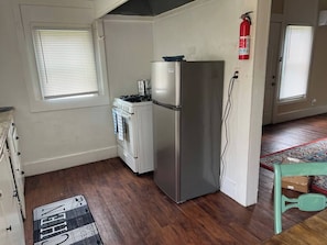 Una cocina con refrigerador, estufa y horno. Hay un extintor de incendios en la pared y una ventana con persianas.