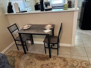 Un comedor con mesa y sillas de madera, una encimera de cocina con electrodomésticos, y una alfombra estampada.