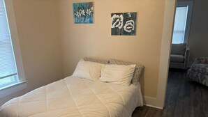 Ein Schlafzimmer mit einem Bett, zwei an der Wand befestigten Bildern, einem Fenster und einer Tür zu einem anderen Raum.