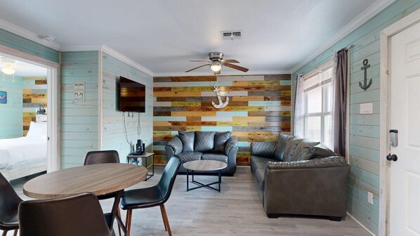 A room with a wooden wall, a ceiling fan, a sofa, a coffee table, and a dining table with chairs. - ACC3 Shared Pool, In Town, Pet Friendly (Port Aransas)