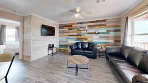 A living room with a flat-screen TV, a ceiling fan, a black leather sofa, a wooden coffee table, and a wooden wall panel. - ACC5 Shared Pool, In Town, Pet Friendly (Port Aransas)