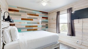 Quarto com cama, ventilador de teto, TV na parede e janela com cortinas.