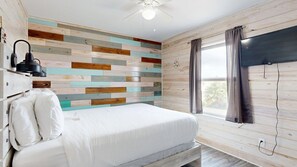 A bedroom with a bed, a ceiling fan, a wall-mounted TV, and a window with curtains. - ACC5 Shared Pool, In Town, Pet Friendly (Port Aransas)