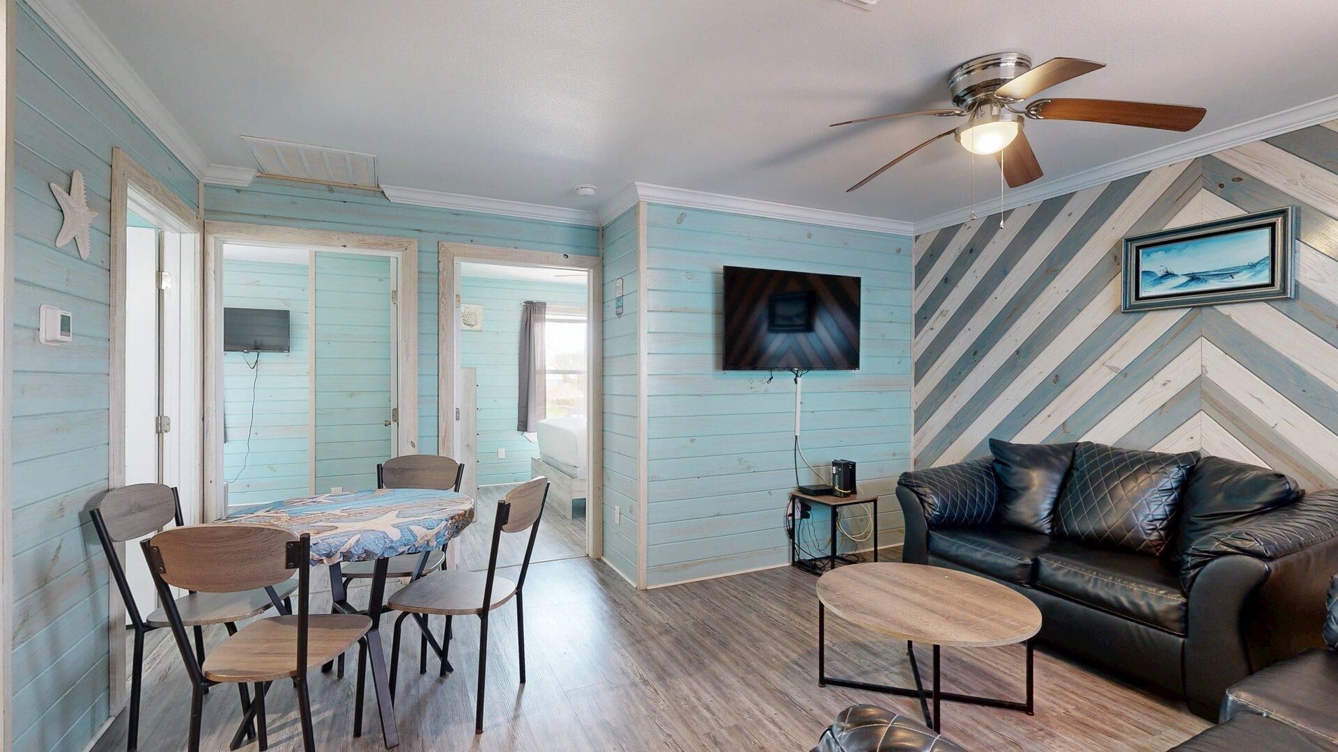 A room with a dining table, chairs, a sofa, a ceiling fan, and a TV mounted on the wall.