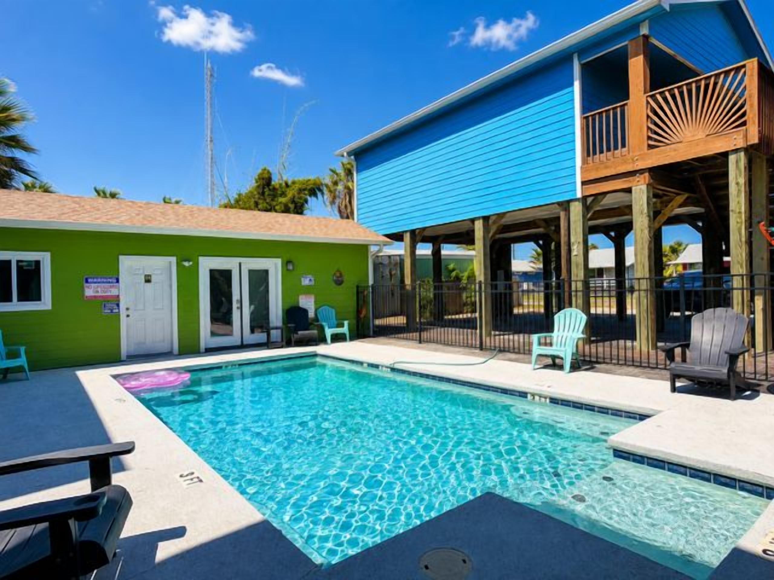 Un área de piscina con una piscina azul cristalina, un espacio de estar cubierto con sillas y un edificio con techo azul.