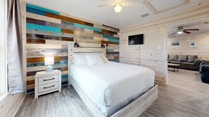 A bedroom with a wooden headboard, a bedside table with a lamp, a flat-screen TV, and a view of the living area. - ACC8 Shared Pool, Pet Friendly, In Town (Port Aransas)