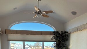 Un comedor con una mesa redonda, sillas, un ventilador de techo y vistas a una zona residencial a través de amplios ventanales.
