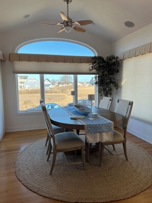Ein Essbereich mit einem runden Tisch, Stühlen, einem Deckenventilator und Blick auf eine Wohngegend durch große Fenster.