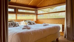 A wooden-paneled room with a large bed, wooden flooring, and a window with blinds. - Peace, Comfort & Nature at This Home (Durbuy)