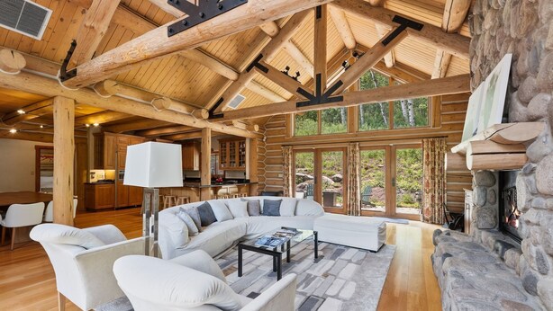 Ein rustikales Blockhaus-Interieur mit Kamin aus Stein, Holzbalkendecke, einem großen, L-förmigen Ecksofa und einem Essbereich.