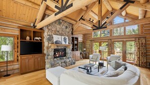 Ein rustikales Blockhaus-Interieur mit Kamin aus Stein, Holzbalken, einem weißen Sofa und einem Fernseher.
