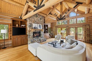 Ein rustikales Blockhaus-Interieur mit Kamin aus Stein, Holzbalken, einem weißen Sofa und einem Fernseher.