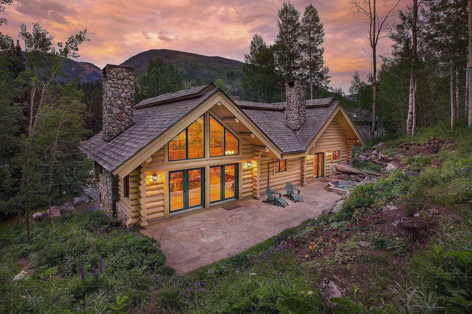 Log cabin with a stone patio, hot tub, and mountain backdrop at twilight.