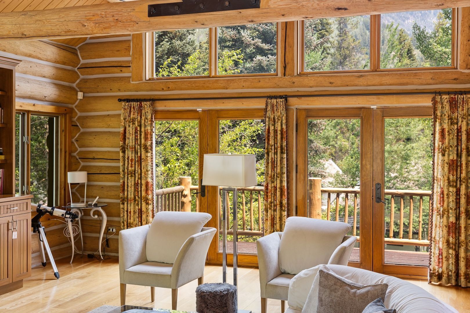 A log cabin interior with a wooden floor, two white armchairs, a small table, a lamp, and a view of the forest through large windows.