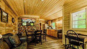 Interior acolhedor de uma cabana com área de refeições, cozinha e uma televisão exibindo 'Serenity-Smokes'.