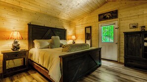 Quarto de cabana de madeira com cama, mesas de cabeceira, cómoda e porta que dá para uma área verde externa.