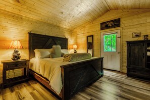 Ein Schlafzimmer einer Holzhütte mit einem Bett, Nachttischen, einer Kommode und einer Tür zu einem grünen Außenbereich.