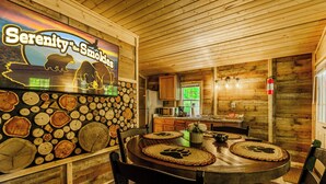 Interior de madeira com mesa de jantar, cadeiras, área de cozinha e uma parede com arte decorada com tema de urso e floresta.