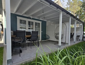 A covered patio with a table and chairs, a hanging chair, and a grill area. - Comfortable Houses With Terraces (Rewal)