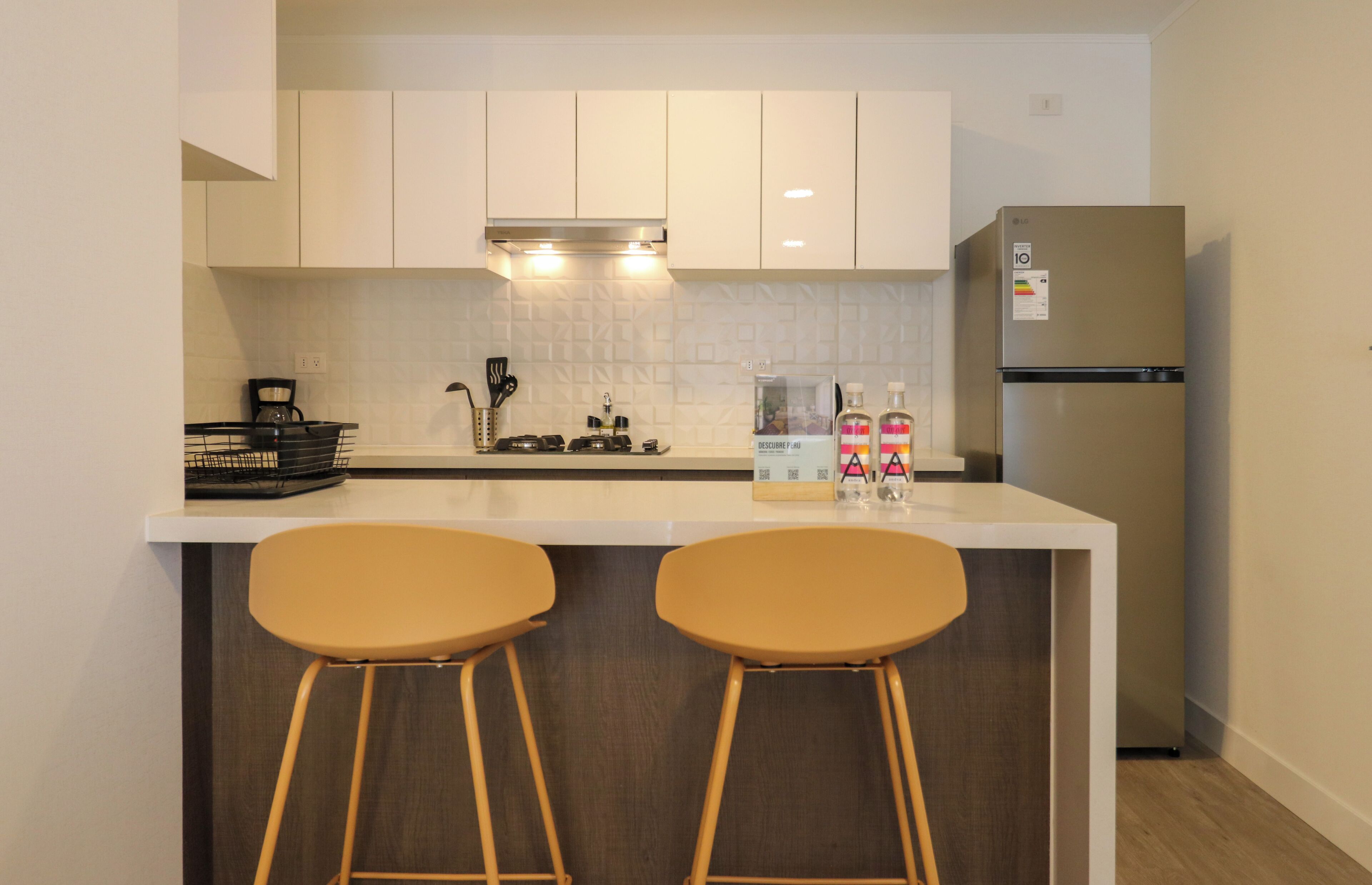 A kitchen with white cabinets, a stainless steel refrigerator, two bar stools, a coffee maker, and a bottle of hand sanitizer.