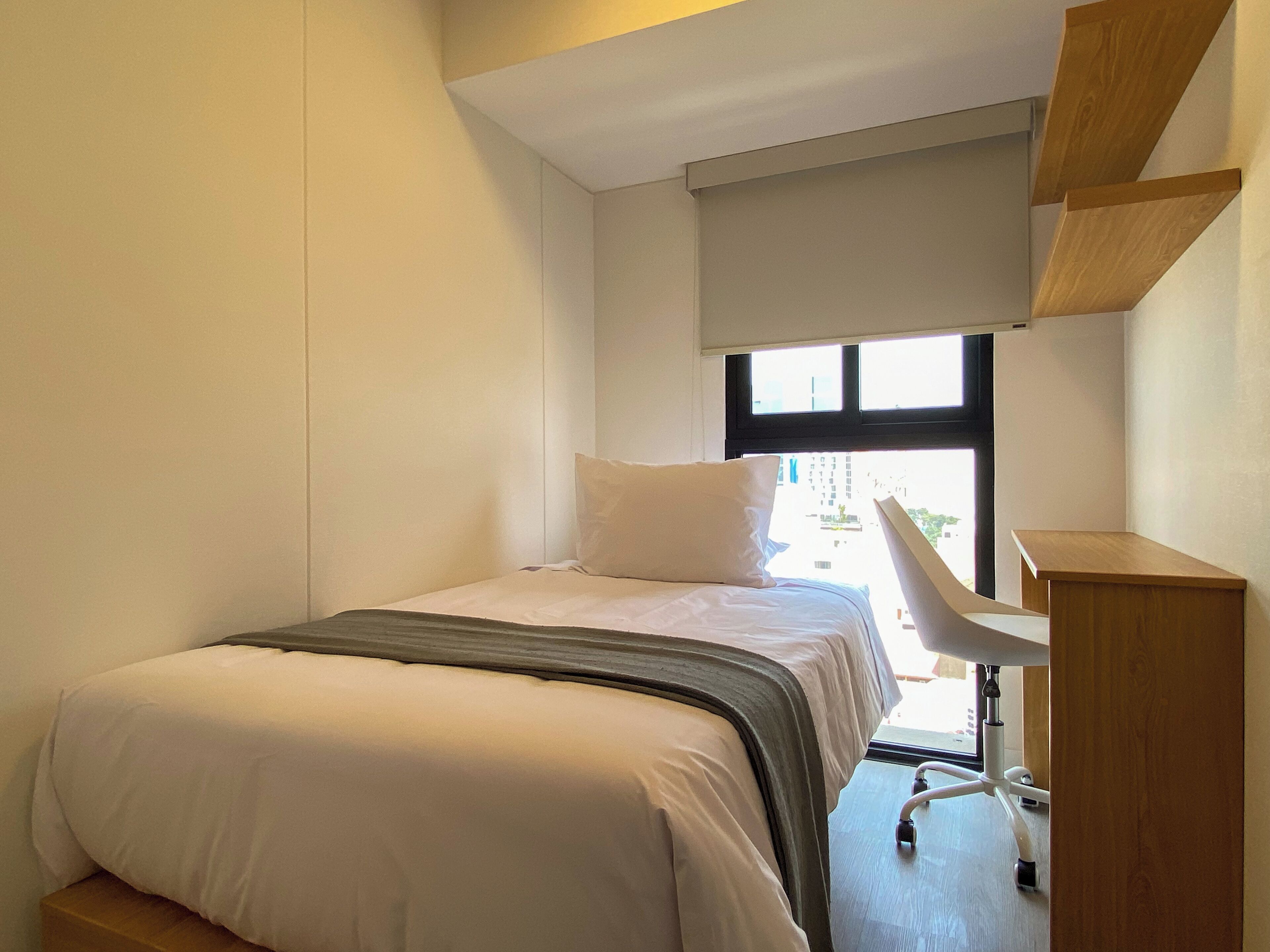 Une petite chambre d’hôtel moderne, équipée d’un lit, d’un bureau et d’une fenêtre.