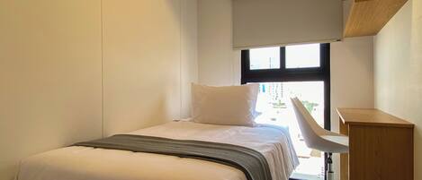 Une petite chambre d’hôtel moderne, équipée d’un lit, d’un bureau et d’une fenêtre.