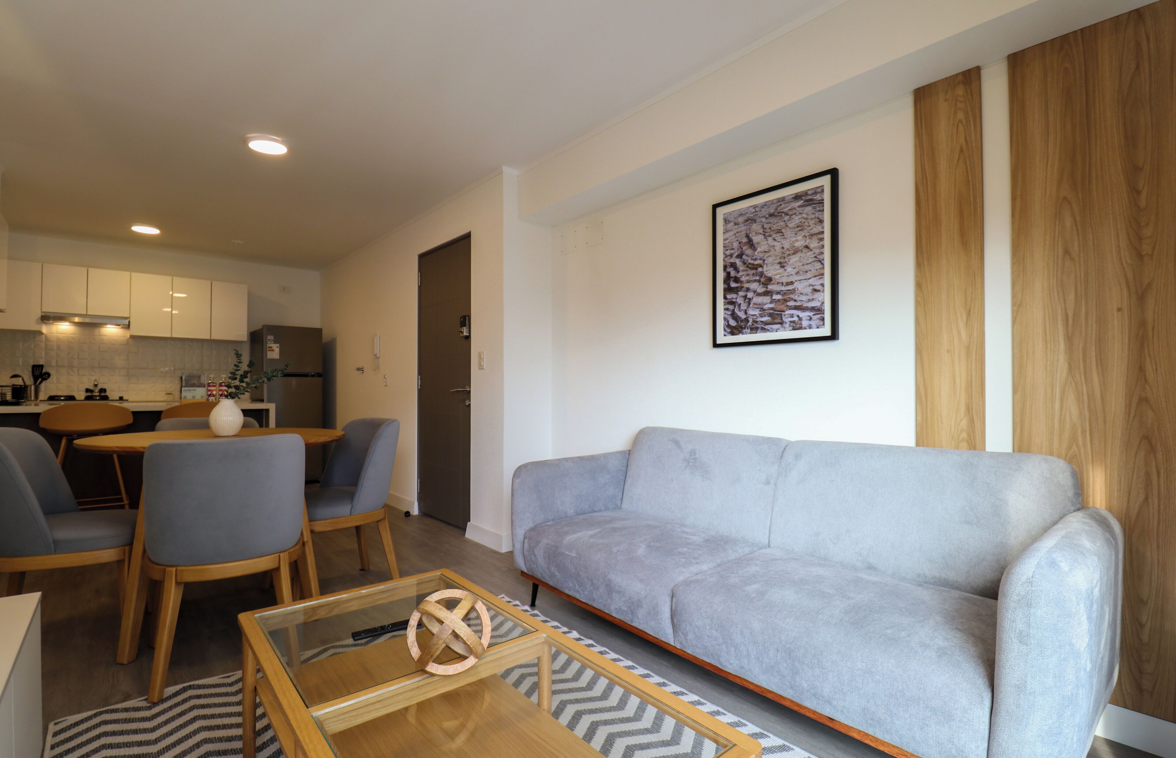 Un salon moderne avec un canapé gris, une table à manger en bois et des chaises. On aperçoit une cuisine à l’arrière-plan, avec des meubles blancs et un réfrigérateur.