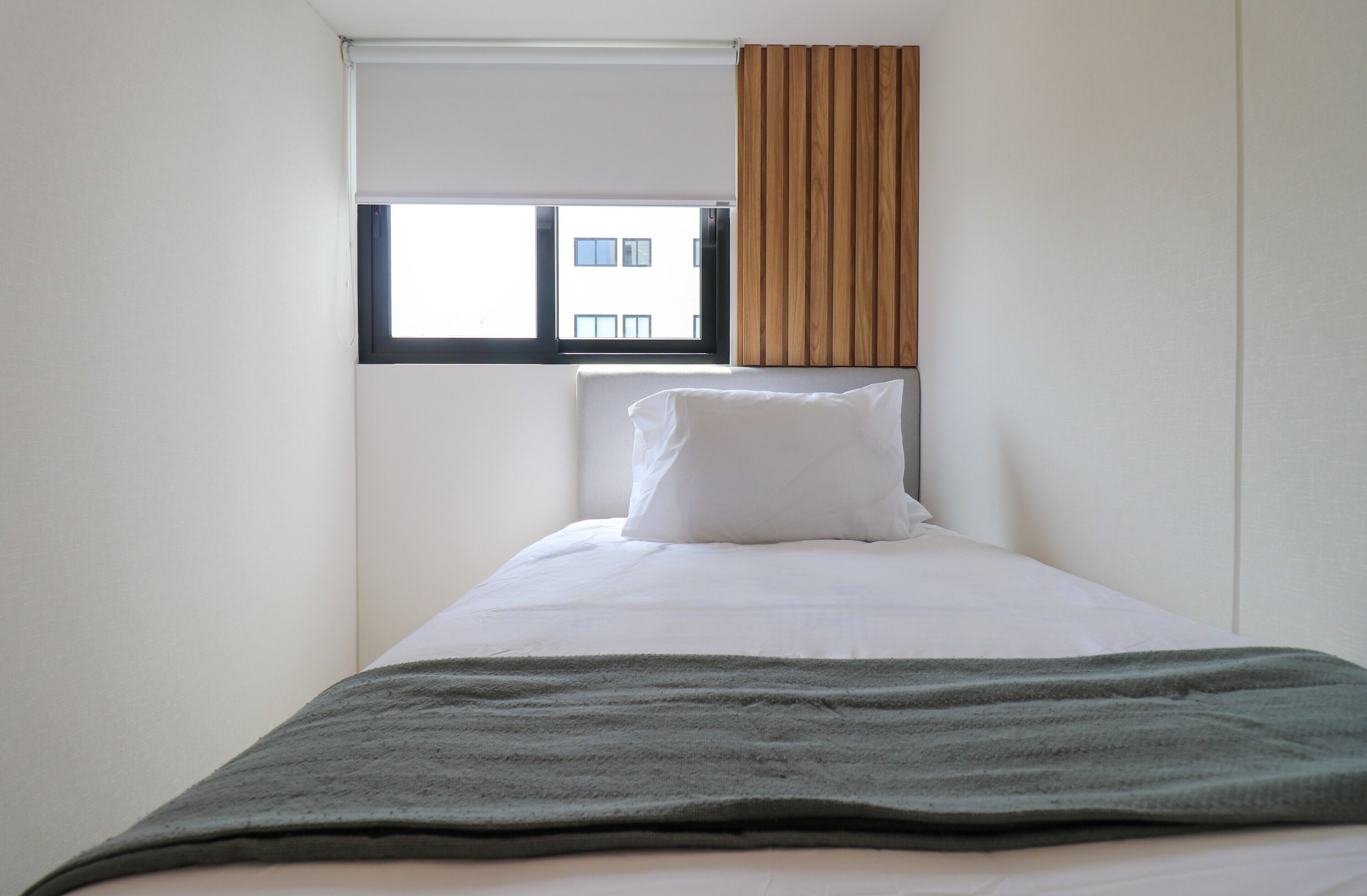 A single bed with a white pillow and a green blanket, a window with blinds, and a view of a building outside.